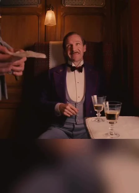 Gustave H. in a purple tuxedo looking surprised as a hand holds out a bill.