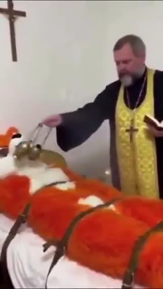 Священник в желтой одежде освящает огромную красную рыбу с крестом на фоне