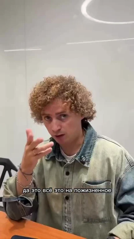 a man with curly hair gesturing while sitting at a desk with Russian text overlay