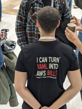 Man wearing a black t-shirt with the text 'I CAN TURN YAML INTO AWS BILLS WHAT'S YOUR SUPERPOWER?'