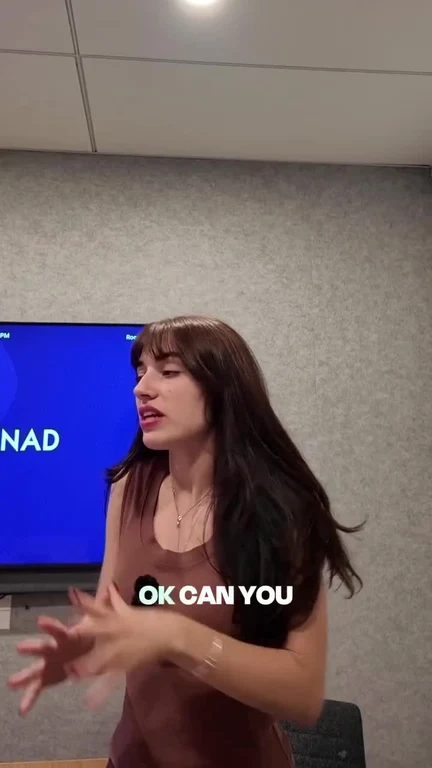 Woman in an office meeting room gesturing while asking 'OK can you' in front of a blue presentation screen