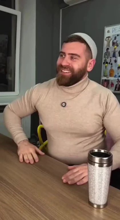 Bearded man in a beige turtleneck laughing while sitting at a desk with a silver travel mug.