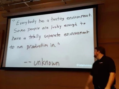A presenter stands beside a projection screen showing a handwritten quote: "Everybody has a testing environment. Some people