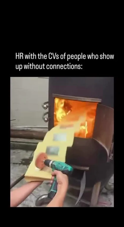 A hand holding a power drill with a sanding pad against a piece of wood, positioned next to a wood-burning stove with