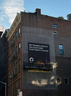 Billboard on a brick building with the text 'WE TRAINED COPILOT ON YOUR CODE. THAT'S WHY IT SUCKS'