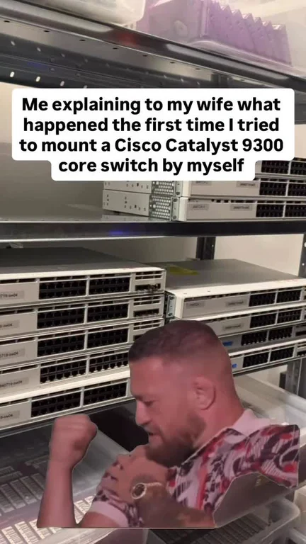 A man flexing his muscles in front of a server rack with network switches, captioned about the difficulty of mounting a Cisco Catalyst 9300 switch by himself.