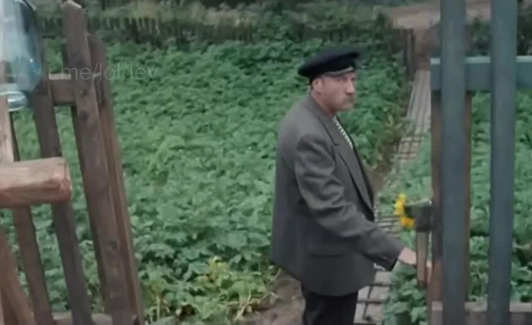 Man in a gray jacket and black beret walking through a garden gate