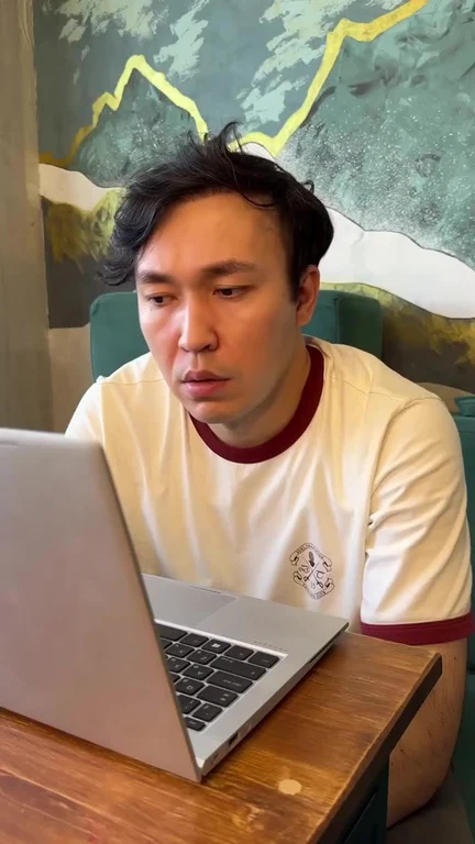 Man in a white t-shirt sitting at a wooden table, staring at a laptop with a flat, disappointed expression