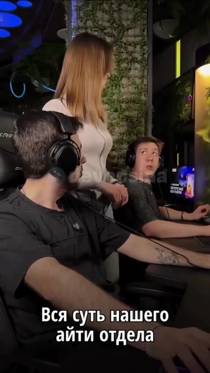 Two men in gaming headsets sitting at computers with a woman standing behind them