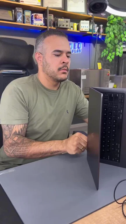 A man with a green t-shirt looking at a laptop standing vertically on a desk mat, surrounded by tech gadgets and plants.