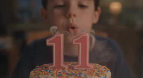 A young boy blowing out pink number 11 candles on a sprinkle-covered birthday cake.