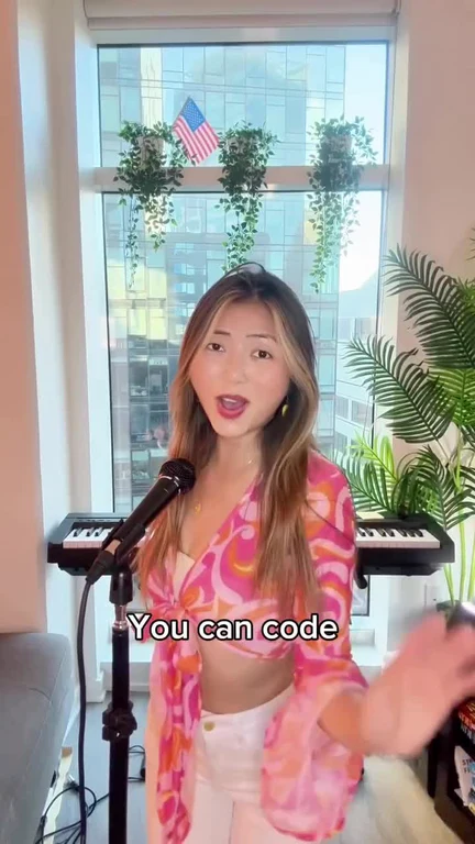 A woman in a bright pink top standing in a modern apartment with a view, microphone, and keyboard, with text overlay reading 'You can code'.