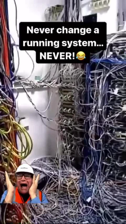 a man in a white hard hat and orange vest reacts in horror to a tangled mess of wires with the caption 'Never change a running system... NEVER!'