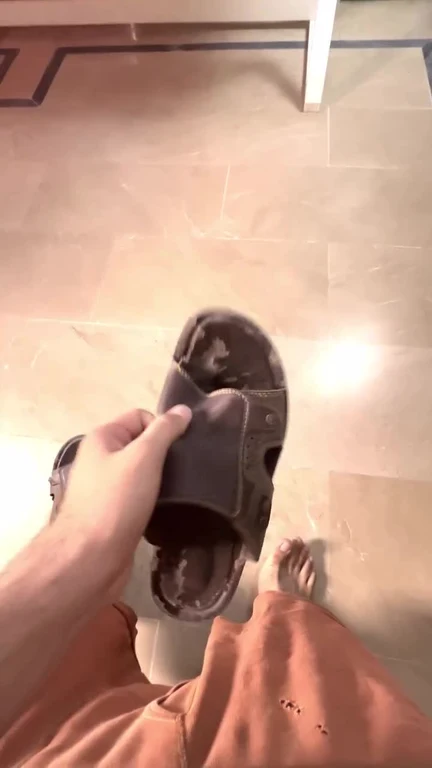 First-person view of a hand holding a worn-out, falling-apart sandal above a bare foot on a tiled floor