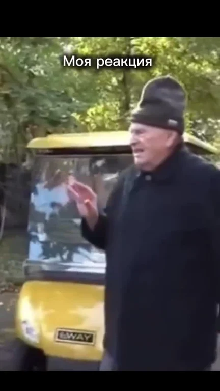 Man in black coat and hat gesturing in front of yellow vehicle with russian text