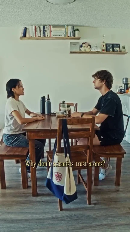 Two people seated across a wooden table with text overlay 'Why don't scientists trust atoms?'