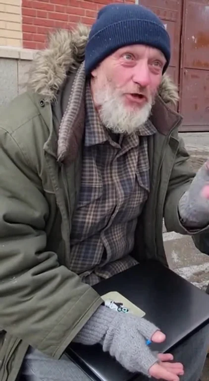 Old homeless man with a white beard and blue beanie sitting on a sidewalk holding a laptop with stickers