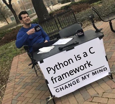 Man sitting at a table with a sign that reads 'Python is a C framework CHANGE MY MIND'