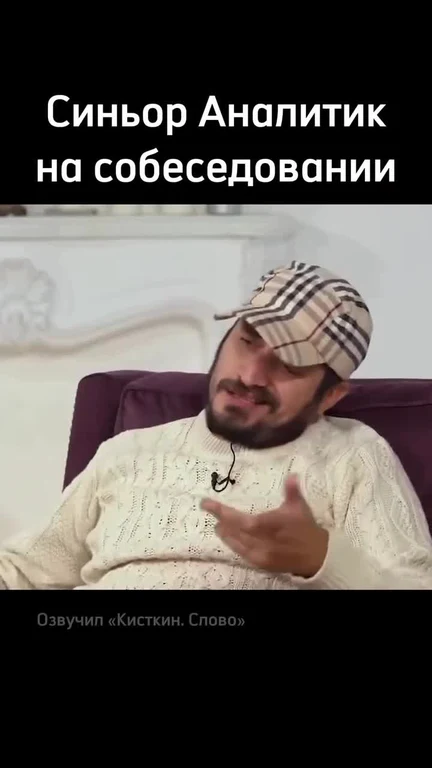 A man in a beige sweater and plaid cap gesturing during a job interview.