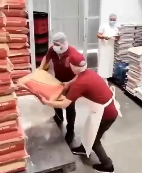 Two workers in red shirts and hairnets struggling to move a heavy bag