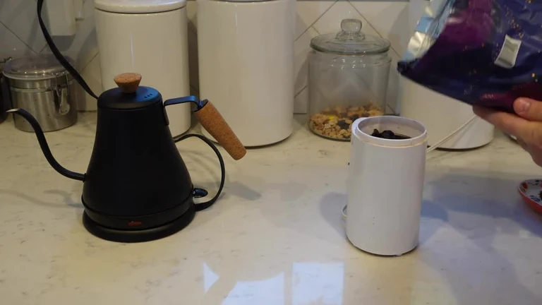 Hand pouring coffee beans from a bag into a white electric coffee grinder next to a black gooseneck kettle on a kitchen counter.