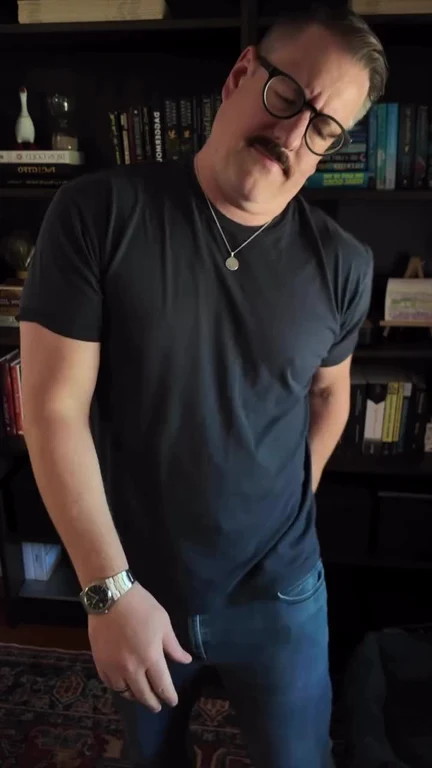 A man with a mustache and glasses shaking his head and grimacing in disappointment in front of a bookshelf.