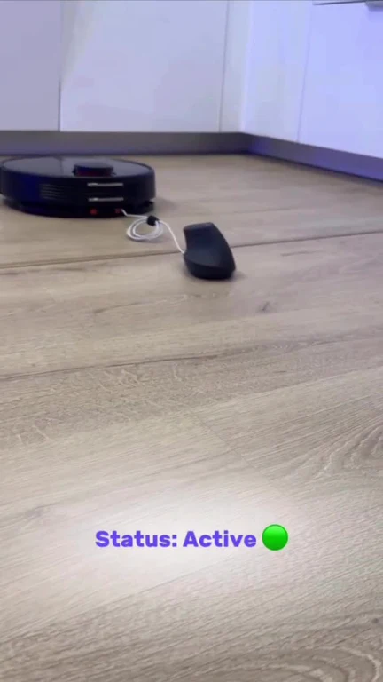 A robot vacuum cleaner dragging a wired computer mouse across a wooden floor with text overlay Status: Active