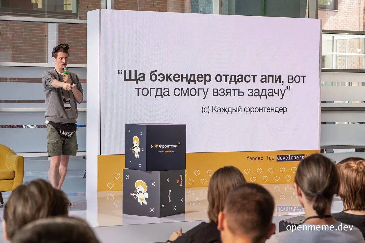 a man in a gray shirt and green shorts speaks into a microphone on a stage with a large screen behind him that displays a quote in Russian, with an audience seated in front of him