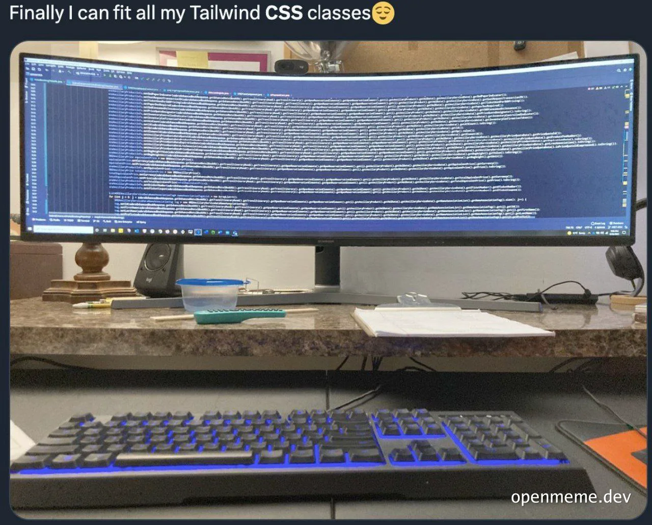 An ultrawide curved monitor displaying one extremely long line of Tailwind CSS classes, with a mechanical keyboard and desk